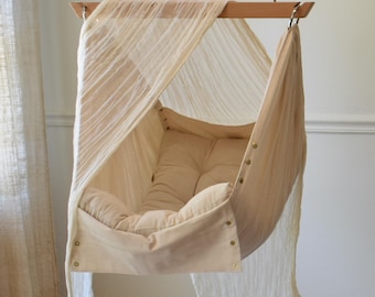 May include: A beige fabric baby swing with a canopy. The swing is suspended from a wooden bar with a chain. The swing has a padded cushion and a soft fabric lining.