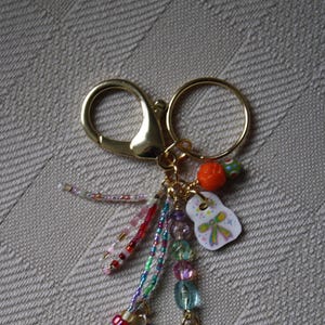 Puede incluir: Un llavero dorado con una variedad de cuentas de colores, dijes y un dije de estrella verde. Los dijes incluyen una pequeña cuenta naranja, un dije blanco con un lazo y un dije en forma de corazón.