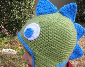 May include: A green and blue crocheted hat with a large eye and a dinosaur-like design. The hat has a long blue tail and is worn by a child.