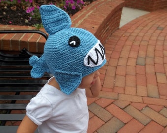 Puede incluir: Un niño con un gorro de tiburón de crochet azul con dientes blancos y un ojo negro. El gorro tiene una aleta a cada lado.