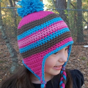 May include: A pink, brown, and blue crocheted hat with a blue pom-pom on top. The hat has ear flaps that tie under the chin.