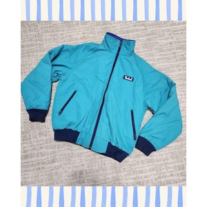 May include: A teal long-sleeve jacket with a high collar and a full-length dark blue zipper. The jacket features dark blue ribbed cuffs and a matching dark blue ribbed hem. A small rectangular patch with the white text Woolrich is on the chest.