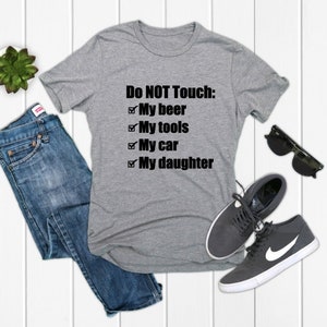 May include: A heather gray t-shirt with the text "Do NOT Touch: My beer, My tools, My car, My daughter." The shirt is paired with blue jeans, sunglasses, and gray sneakers.