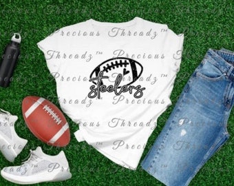 Peut inclure: T-shirt blanc avec un graphique de football noir et le texte "Steelers". Le t-shirt est posé sur une surface herbeuse verte avec une paire de baskets blanches, une bouteille d'eau et un jean bleu.