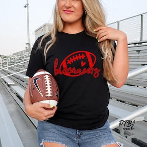 Puede incluir: Camiseta negra con un diseño de balón de fútbol rojo y la palabra "buccaneers" en escritura cursiva. Un balón de fútbol marrón y blanco se sostiene frente a la camiseta. La camiseta se combina con vaqueros azules rotos.