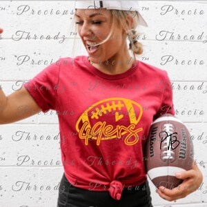 May include: A red t-shirt with a yellow and gold football graphic that says "49ers". The football has a heart on it. The shirt is being held by a person holding a brown football.