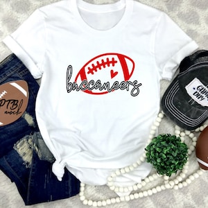 May include: White t-shirt with a red and black football graphic that says "buccaneers". The shirt is paired with blue jeans, a black hat with "Game Day" text, and a decorative football.