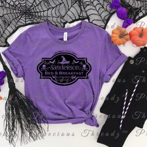 Puede incluir: Camiseta morada con un gráfico negro de un letrero que dice "Sanderson Bed & Breakfast Est. 1693" con un sombrero de bruja, murciélagos y calaveras.