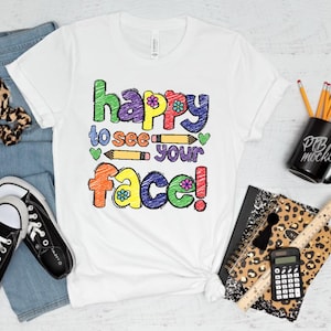 May include: White t-shirt with the text "happy to see your face" in colourful, hand-drawn lettering. The image includes a pair of black trainers, blue jeans, and a black mug with pencils.