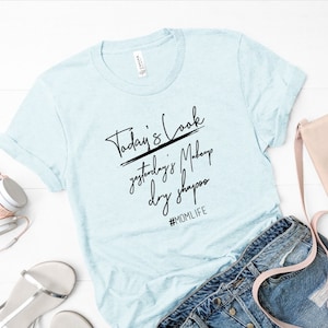 May include: A light blue t-shirt with the text "Today's Look yesterday's Makeup dry shampoo #MOMLIFE" printed in black.