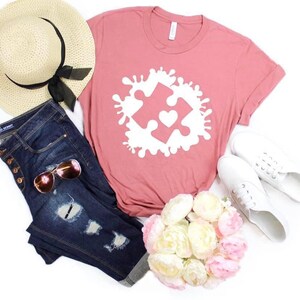 May include: A pink t-shirt with a white puzzle piece design featuring a heart in the center. The puzzle piece is surrounded by white paint splatters. The shirt is paired with blue jeans, a straw hat, white sneakers, and a bouquet of white and pink flowers.