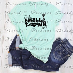 May include: A light blue t-shirt with a black graphic that says "Just a Small Town Girl" with a silhouette of North Carolina and a windmill. The shirt is on a white surface with a pair of blue jeans.