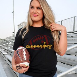 May include: Black t-shirt with a football graphic and the word "commanders" in yellow script. A brown and white football is held in the hand. The shirt design features a football shape with a heart.
