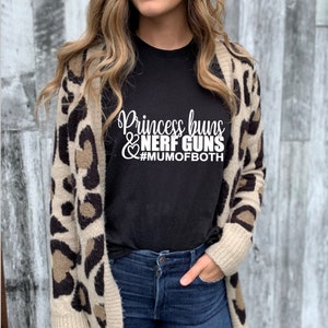 May include: A black t-shirt with white text that reads "Princess buns & NERF GUNS #MUMOFBOTH". The person wearing the shirt is also wearing a brown and black leopard print cardigan and blue jeans.