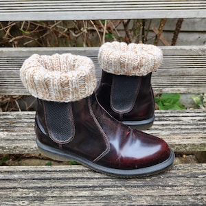 May include: A pair of dark burgundy leather Chelsea boots featuring black elastic side panels and thick, ribbed-knit beige and white yarn cuffs that fold over the top edge of the ankle, creating a textured contrast against the smooth, polished leather.