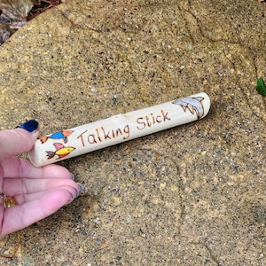 May include: A cylindrical wooden talking stick featuring wood-burned text that reads Talking Stick. The surface is decorated with a wood-burned and coloured illustration of a dolphin on the right side and two small colourful fish on the left side.