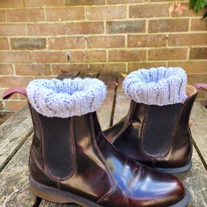 May include: A pair of dark burgundy leather Chelsea boots featuring a glossy finish, black elastic side panels, and a pull tab at the heel. Each boot is topped with a light blue, chunky knit ribbed cuff that extends slightly above the collar.