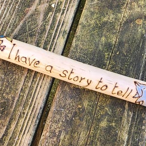 May include: A wooden stick with the text "I have a story to tell" burned into the surface. The stick is decorated with small drawings of fish and a dolphin. The background is a weathered wooden surface.