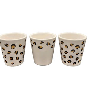 May include: Three white ceramic cups featuring a repeating leopard print pattern in black and gold. The cups have a tapered cylindrical shape with a smooth finish. The two outer cups are fully covered in the pattern, while the centre cup is plain.