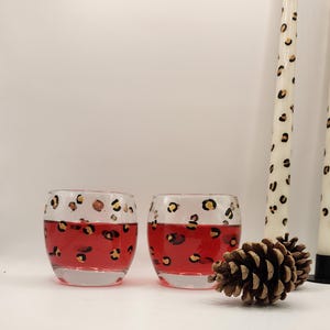 May include: Two clear glass votive candle holders filled with red wax, each featuring a hand-painted black and gold leopard print pattern around the rim. A brown pinecone sits in front of the glasses, next to two tall white taper candles.