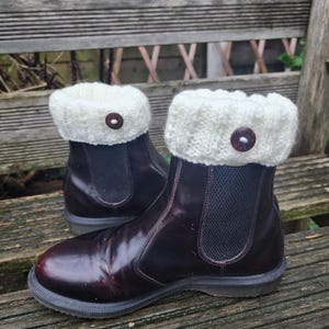 May include: Burgundy Chelsea boots with cream knitted boot cuffs, each fastened with a brown button. The boots are displayed on a wooden bench. The cuffs add a decorative touch.
