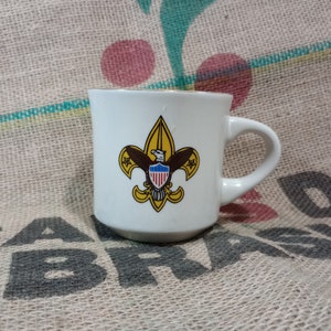 Könnte beinhalten: Weiße Keramiktasse mit goldfarbenem Rand und einem gelben Boy Scout-Emblem mit einem Adler und der amerikanischen Flagge.