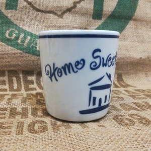 Puede incluir: Taza de cerámica blanca con un borde azul y letras azules que dicen "Home Sweet Home" con una ilustración estilizada de una casa.