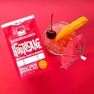 May include: A red single-serve cocktail mix packet with the text "Leisuremann's Hurricane Tropical Fruit & Hibiscus Single Serve Cocktail Mix Net Wt. 0.42oz (12g)". A glass with a red cocktail, a cherry, and an orange slice is in the background.