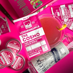 May include: A single-serve cocktail mix sachet for making a Cosmo. The sachet is pink and white with the text "Leisuremann's The Cosmo Tart Berry & Sweet Citrus Syrup" and "Single Serve Cocktail Mix Net Wt 0.21oz (6g)". The sachet is on a pink surface with a pink cocktail glass, a bottle of vodka, and several small round containers with cocktail mix.