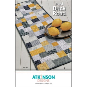 May include: A quilting pattern for a table runner called "Mini Brick Road" by Atkinson Designs. The pattern features a patchwork design with yellow, gray, and black squares. The pattern is shown on a table runner with lemons on a plate.