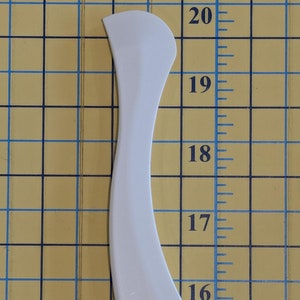 Puede incluir: Una herramienta de plástico blanco y curvada, con un extremo redondeado y un agujero en la parte inferior. La herramienta mide aproximadamente 20 cm de largo y está colocada verticalmente sobre un fondo de cuadrícula amarilla con líneas azules y marcas en cm.