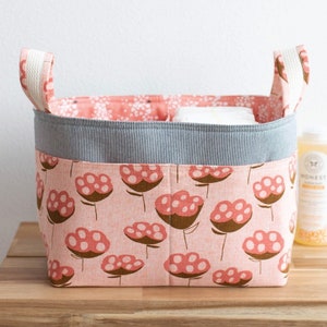 May include: A pink fabric basket with white handles and a divided lining. The basket has a floral print with brown stems and pink flowers with white dots. The basket is sitting on a wooden table.