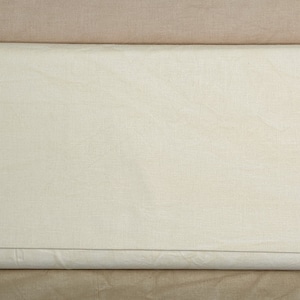 May include: A roll of off-white fabric, tightly wound and secured with white string. The fabric has a subtle texture and is layered over a tan fabric. The image showcases the fabric's color and weave.