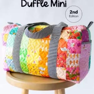 May include: A quilted patchwork duffle bag featuring a colorful array of fabric squares in shades of pink, orange, yellow, green, and blue. The bag has black and white striped handles and side accents. Text reads Patchwork Duffle Mini 2nd Edition.