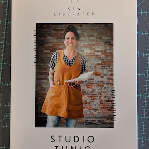 May include: A book cover with the text "SEW LIBERATED" and "STUDIO TUNIC". The cover features a woman wearing a brown apron with pockets, holding a notebook. The background is a brick wall.