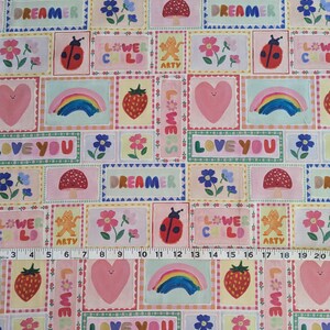以下が含まれることがあります： ピンクと白の生地に、カラフルな正方形が繰り返しパターンで描かれています。各正方形には、ハート、虹、テントウムシ、キノコ、花、そして「Love You」、「Dreamer」、「Flower Child」、「We Are Artsy」というテキストなど、さまざまなデザインが施されています。