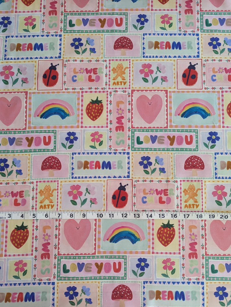 以下が含まれることがあります： ピンクと白の生地に、カラフルな正方形が繰り返しパターンで描かれています。各正方形には、ハート、虹、テントウムシ、キノコ、花、そして「Love You」、「Dreamer」、「Flower Child」、「We Are Artsy」というテキストなど、さまざまなデザインが施されています。