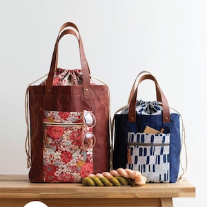 Könnte beinhalten: Zwei Kordel-Tragetaschen, eine mit einem Blumenmuster und eine mit einem blau-weißen geometrischen Muster. Die Taschen stehen auf einer Holzbank mit Garn davor. Der Text "FIREFLY TOTE" und "a drawstring tote in two sizes" befindet sich oben im Bild. Der Text "2 sizes to sew" befindet sich in einem Kreis unten links im Bild. Der Text "noodlehead" befindet sich unten rechts im Bild.