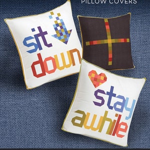 May include: Three pillow covers with a white background and colorful pixelated text. The first pillow cover says "sit down", the second says "stay", and the third says "awhile". The pillow covers are on a blue fabric background. The text "SIT DOWN STAY AWHILE PILLOW COVERS" is at the top of the image. The text "ROBIN PICKENS QUILT PATTERNS" is at the bottom of the image.