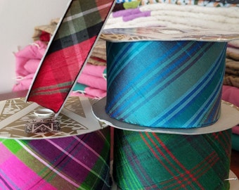 May include: Three spools of plaid ribbon in teal, green, and pink. The ribbon is on a white surface with a gold decorative design.