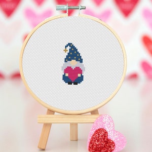 May include: A cross-stitch embroidery hoop with a blue and white gnome holding a red heart. The hoop is on a wooden easel and a pink glitter heart is in the foreground.