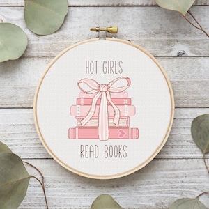 Puede incluir: Un aro de bordado rosa y blanco con el texto "HOT GIRLS READ BOOKS" y una pila de libros rosas con un lazo rosa en la parte superior.