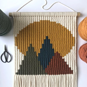 May include: A macrame wall hanging with a geometric design in shades of mustard yellow, forest green, teal blue, and burnt orange. The hanging has a fringe border and is made with natural cotton cord.