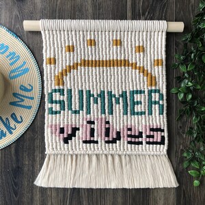 May include: A white macrame wall hanging with the words "Summer Vibes" in colorful yarn. The hanging has a fringe border.