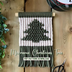 May include: A macrame Christmas tree wall hanging with a light pink and olive green design. The tree is centered on a light pink background, with olive green borders and fringe. The text "Macrame Christmas Tree PDF Pattern Download" is displayed.