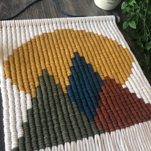 May include: A wall hanging made of woven yarn in a geometric pattern. The pattern features a series of triangles in shades of mustard yellow, forest green, teal blue, and rust brown. The hanging has a fringe of white yarn.