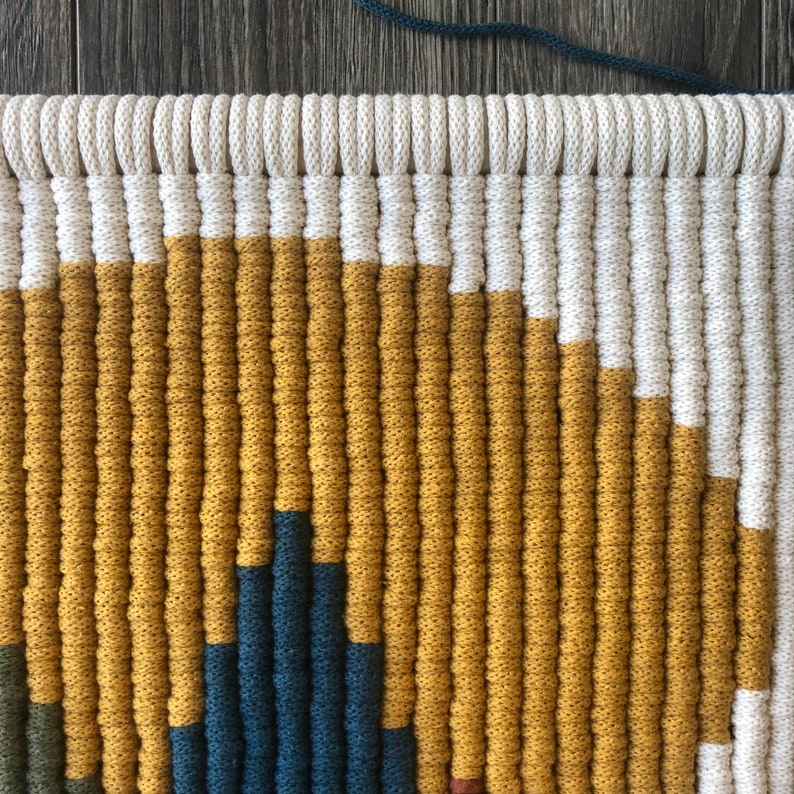 May include: Close-up of a woven wall hanging with a geometric pattern in shades of mustard yellow, teal blue, and white.