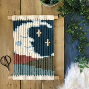 Pode incluir: Uma tape&ccedil;aria de macram&eacute; para pendurar na parede com um design geom&eacute;trico em tons de azul, verde, branco e marrom. O design apresenta duas pequenas cruzes brancas.