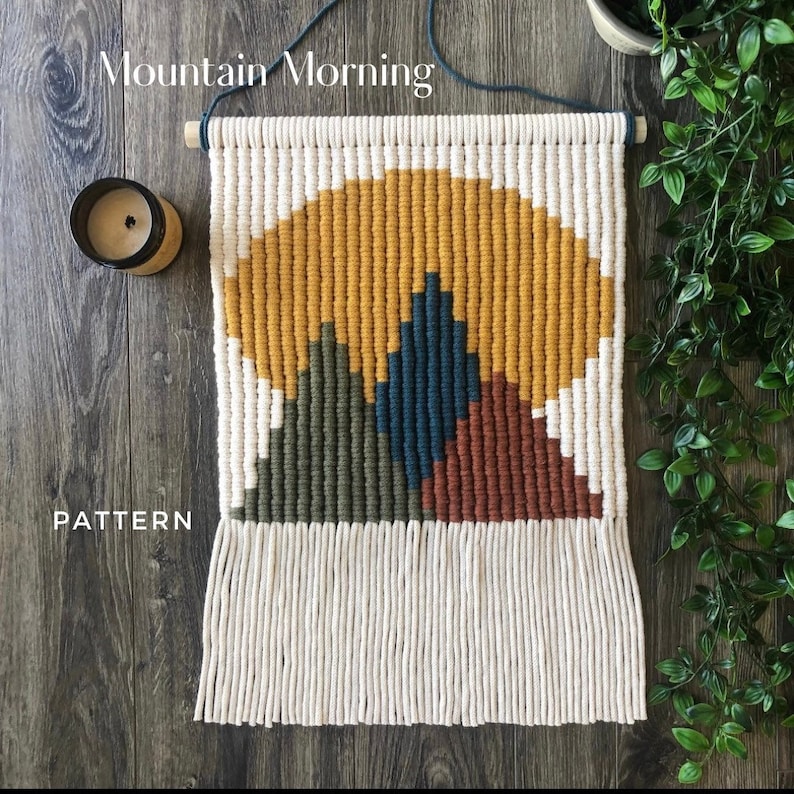 May include: A macrame wall hanging with a geometric design featuring a yellow sun, green, blue, and brown mountains. The wall hanging has a white fringe.