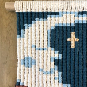 Pode incluir: Uma tape&ccedil;aria de parede feita de fio tecido em tons de creme, azul e azul-petr&oacute;leo. O fio &eacute; tecido em um padr&atilde;o geom&eacute;trico com um sinal de mais dourado no centro.